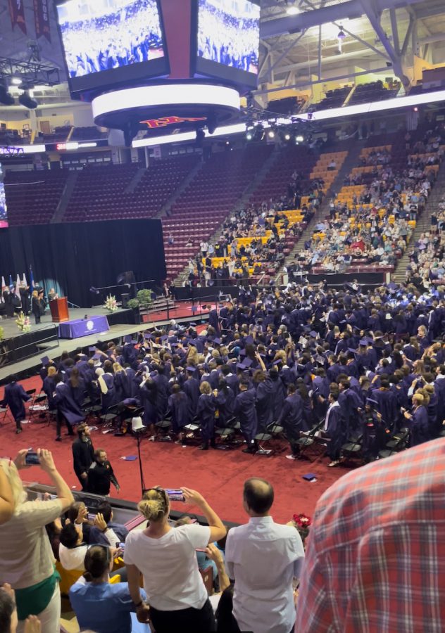 Seniors in black robes toss their tassled hats into their air on the floor of a stadium.