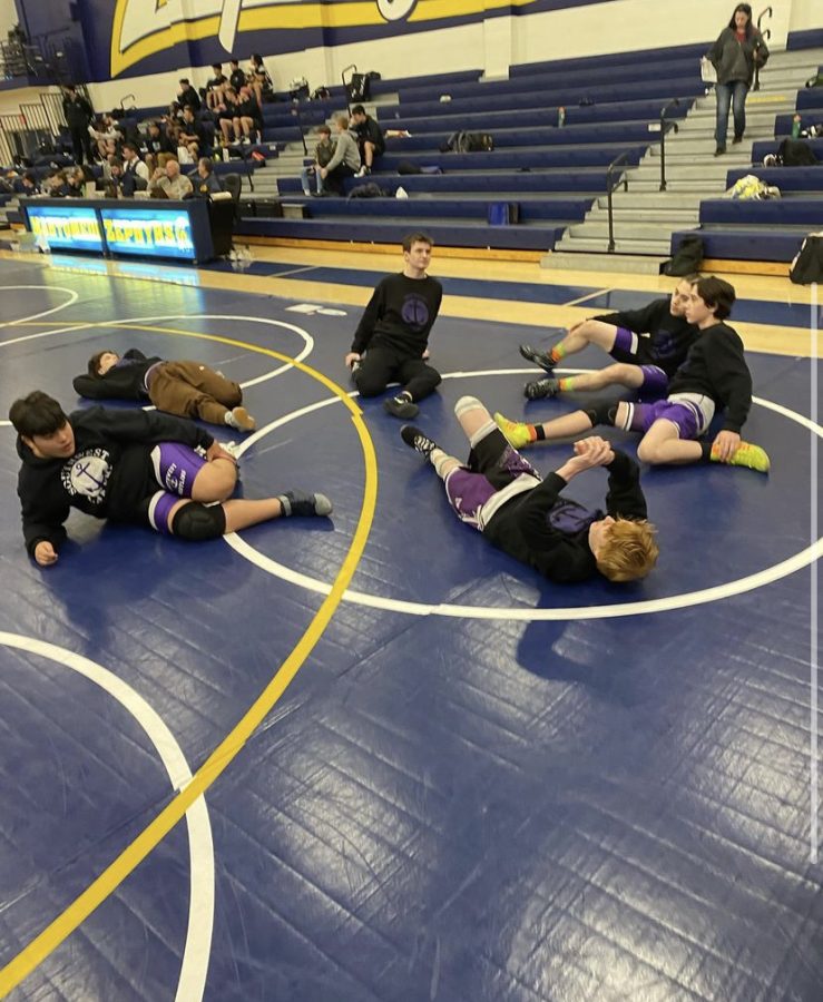 Six students stretch on a purple mat in the foreground. The background shows attendees in the bleachers.